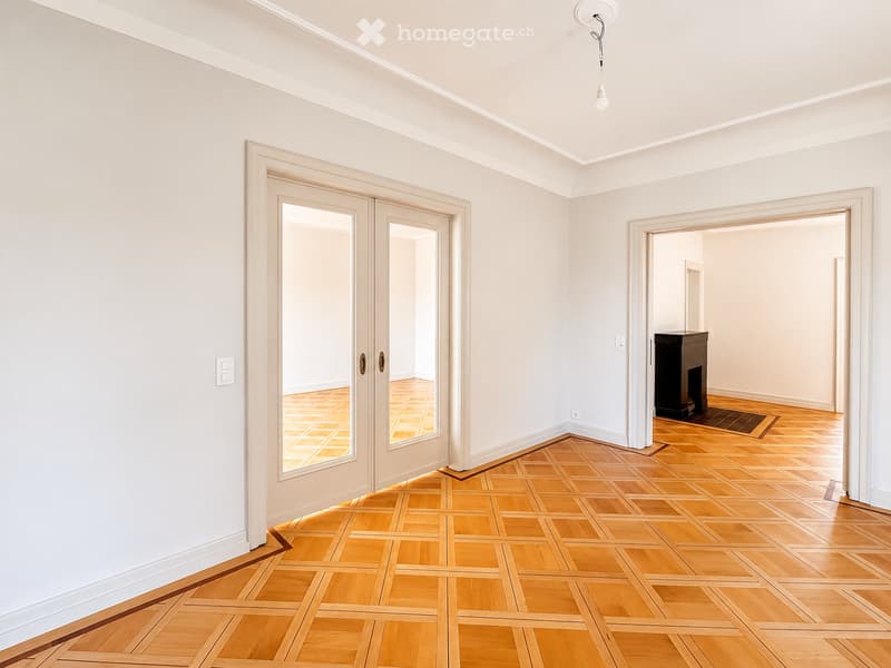 Empty room with light-colored walls, wooden parquet flooring, and a large glass door leading to another space. No furniture or equipment is present.