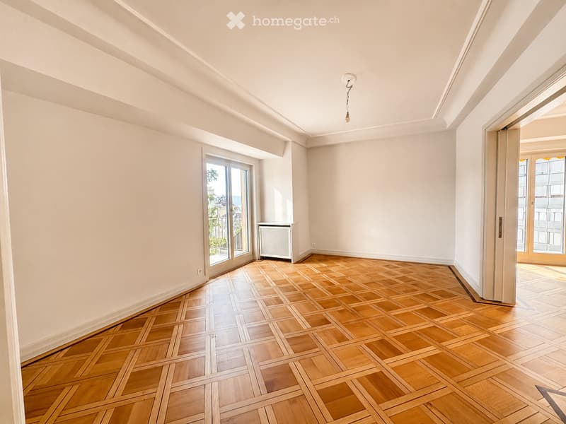 Empty room with wooden herringbone flooring, white walls, large windows, and a radiator. No furniture or personal items present.