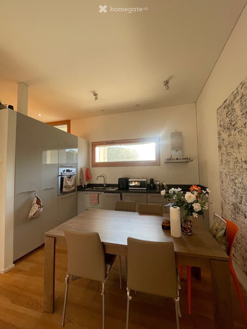 Modern kitchen with light-colored cabinets, a central dining table with chairs, and various appliances including a refrigerator, oven, microwave, and coffee maker. A vase with flowers is placed on the counter.
