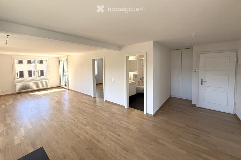 Empty room with wooden floors, white walls, large windows, and a radiator. There are doors leading to a bathroom and a bedroom.