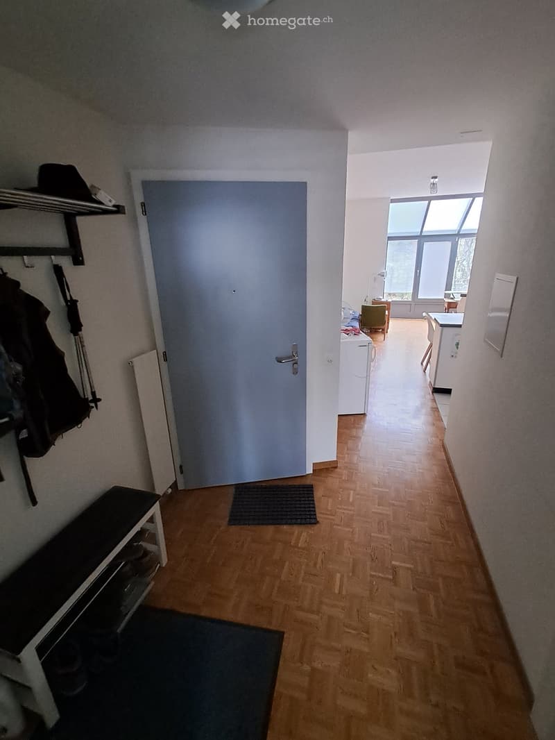 A narrow hallway with wooden flooring, a small bench, and a coat rack. The hallway leads to a room with a window and a kitchen area visible in the background.