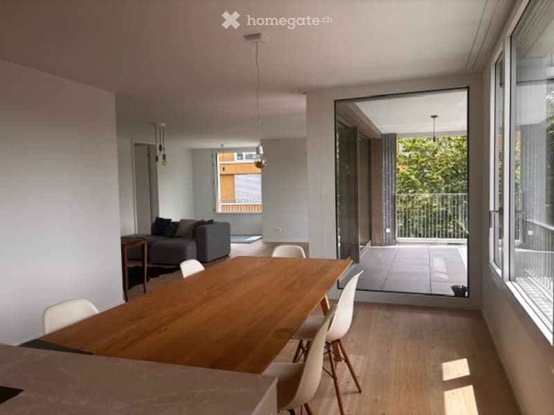 Bright, open-plan living and dining area with large windows and a sliding glass door leading to a balcony. Features a grey sofa, wooden dining table with white chairs, and pendant lighting.