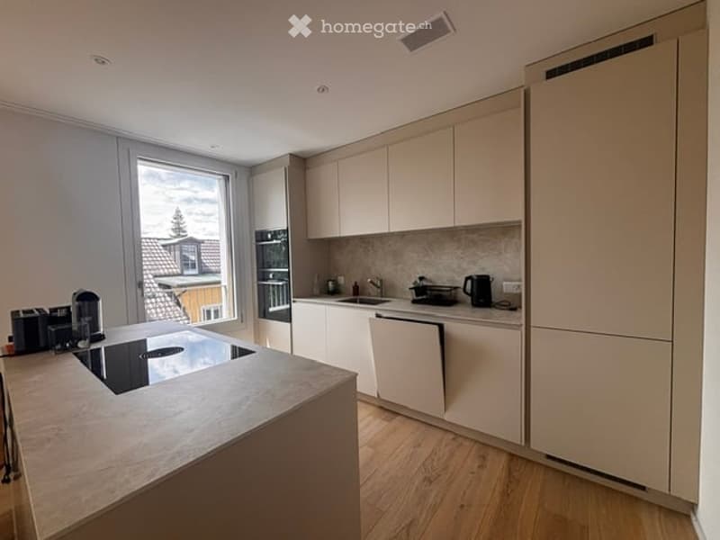 Modern kitchen with light-colored cabinets, a central island, and a large window offering a view of the outside. Appliances include a fridge, oven, and coffee maker.