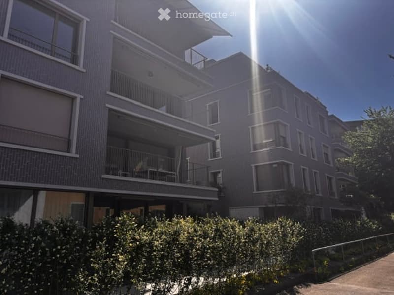 Modern multi-story apartment building with balconies, large windows, and a well-maintained garden area in front.