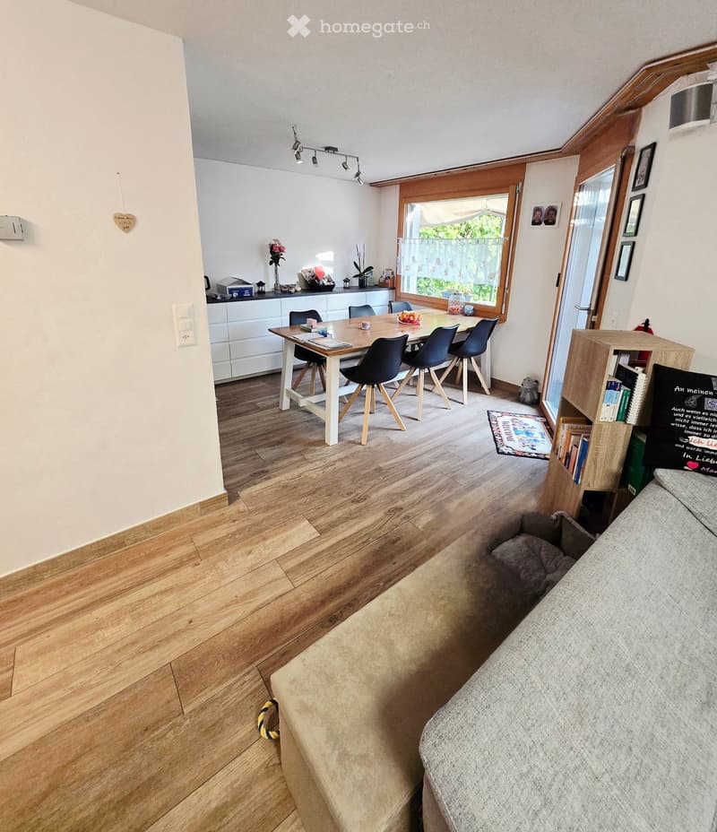 Furnished Room. A modern living and dining area with a grey sofa, a wooden dining table with chairs, a kitchen cabinet, and a bookshelf. The room features wooden flooring and large windows with a view of greenery.