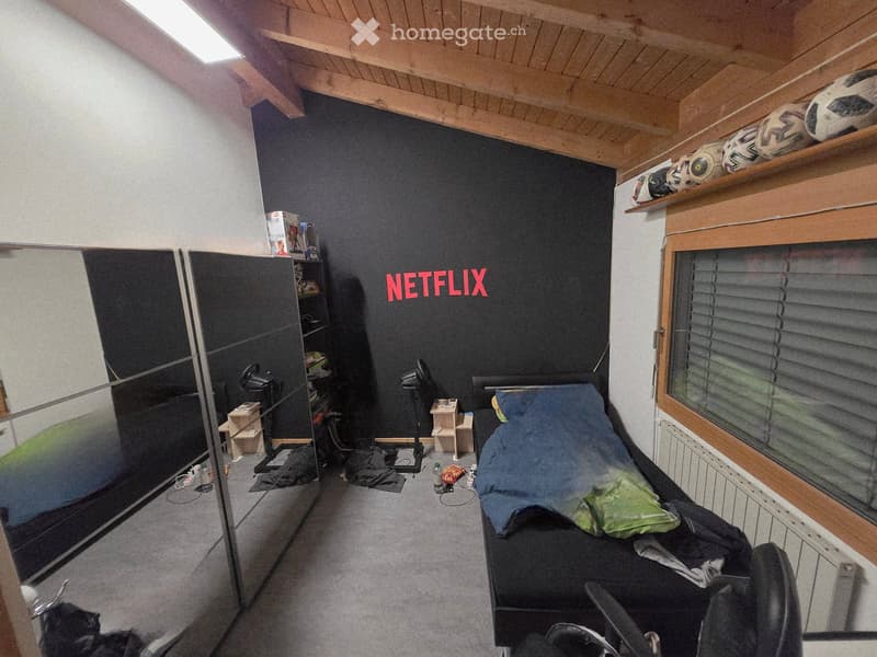 Bedroom. A modern bedroom with a bed, a chair, a shelf, a laptop, and several balls on the shelf. The room features a Netflix logo on the wall and a large window with blinds.
