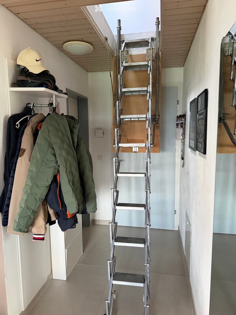 A narrow hallway with a retractable metal staircase leading upwards, coat rack with jackets, hat, and shelves, and framed artwork on the wall.