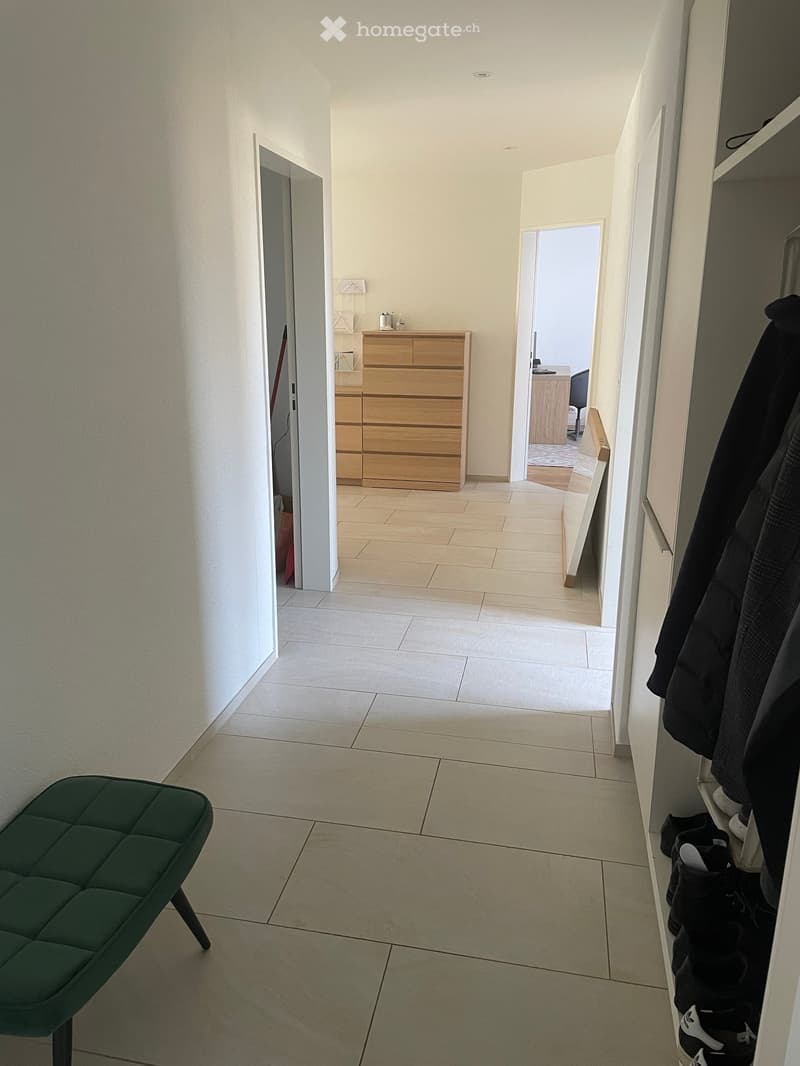 Hallway with tiled floor, green stool, wooden cabinet, and coat rack with jackets and shoes.