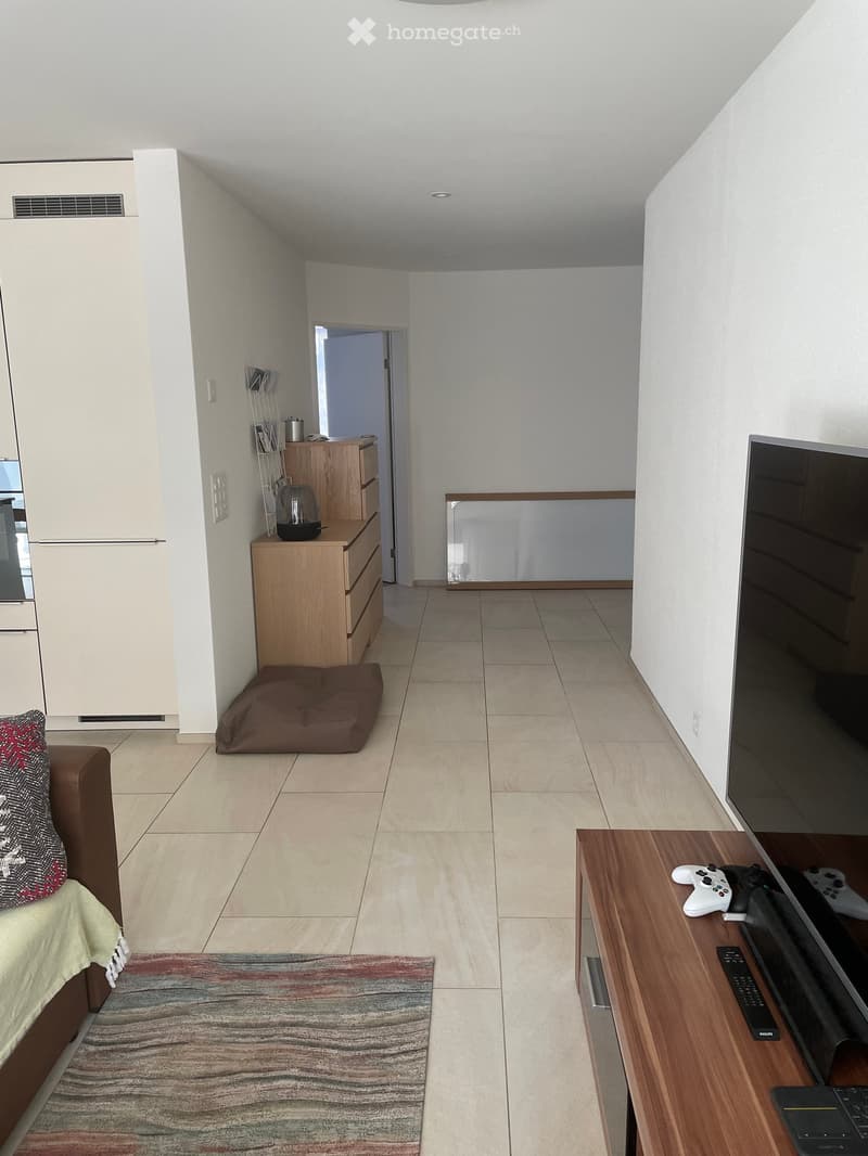 Living room with a sofa, a wooden TV stand, a laptop, remote controls, gaming consoles, a dog bed, a mirror, and a small wooden drawer unit.