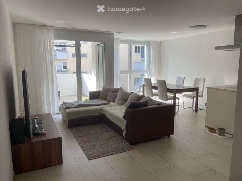 Modern living room with a sectional sofa, coffee table, dining table, chairs, and a TV stand. Large windows and sliding glass doors lead to a balcony.