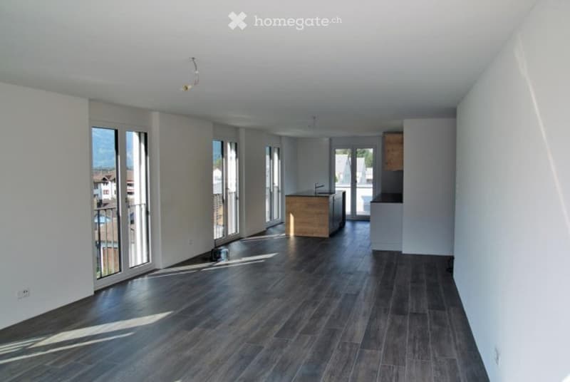 Empty room with dark wooden flooring, white walls, large windows, and a small kitchen area visible in the background.