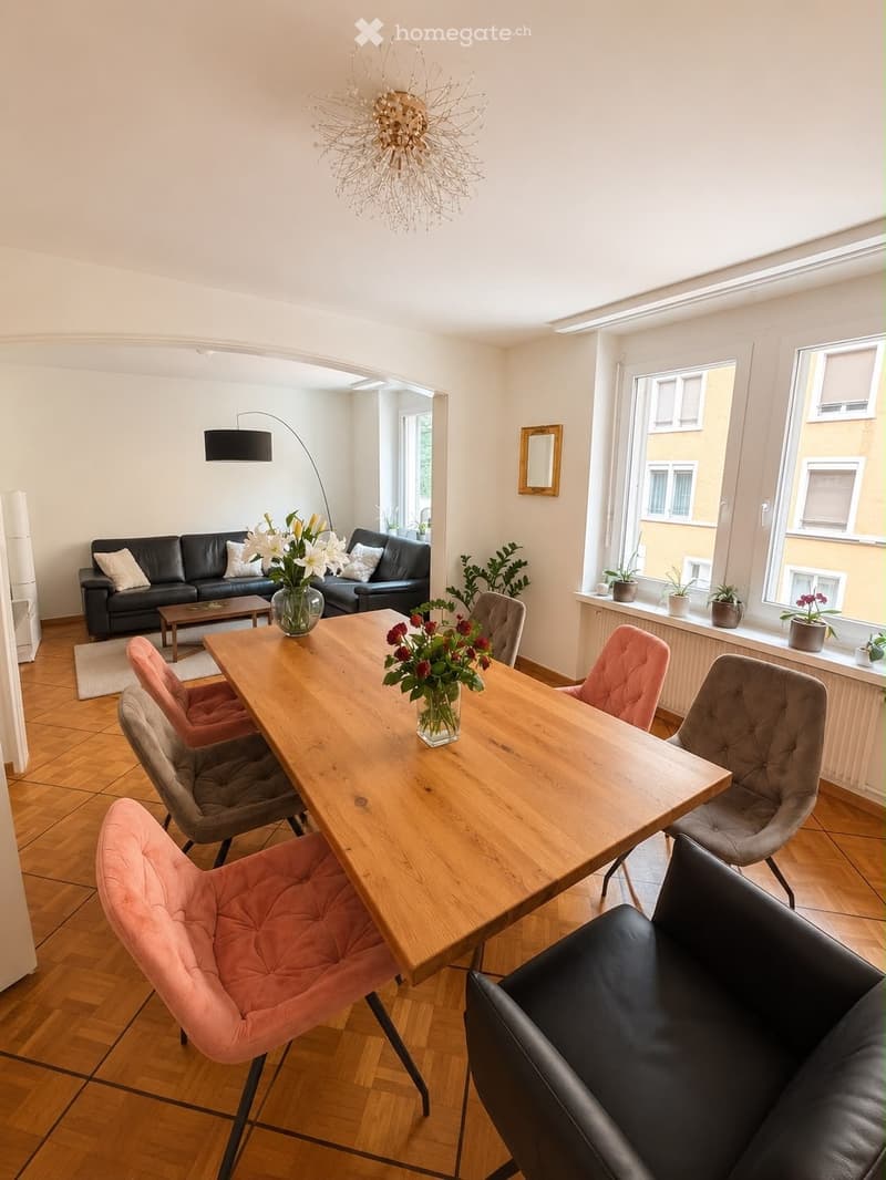 Bright living and dining area with wooden flooring, large windows, a black leather sofa, a wooden coffee table, a dining table with chairs, and decorative vases.