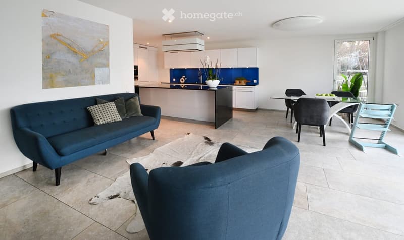 Furnished Room. Modern living and dining area with a blue sofa, armchair, coffee table, dining table with chairs, and a kitchen area with white cabinets and a blue backsplash.