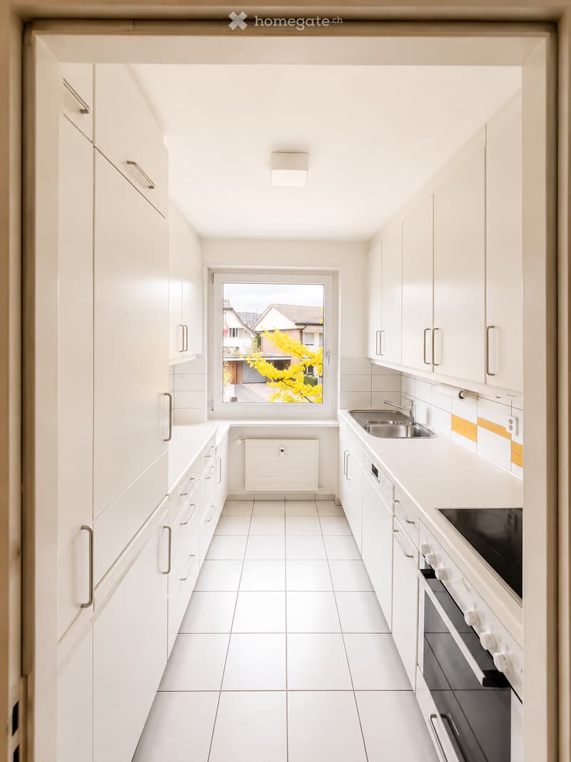 Modern, minimalist kitchen with white cabinets, stainless steel sink, and built-in oven. Features a large window with a view of the street and yellow foliage.