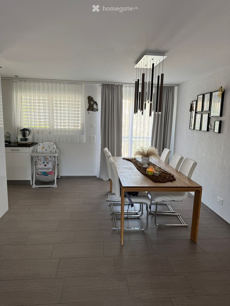 Dining room with a wooden dining table, six white chairs, a baby chair, a kitchen cabinet with a blender, and a chandelier hanging from the ceiling. The room features large windows with curtains and several picture frames on the wall.