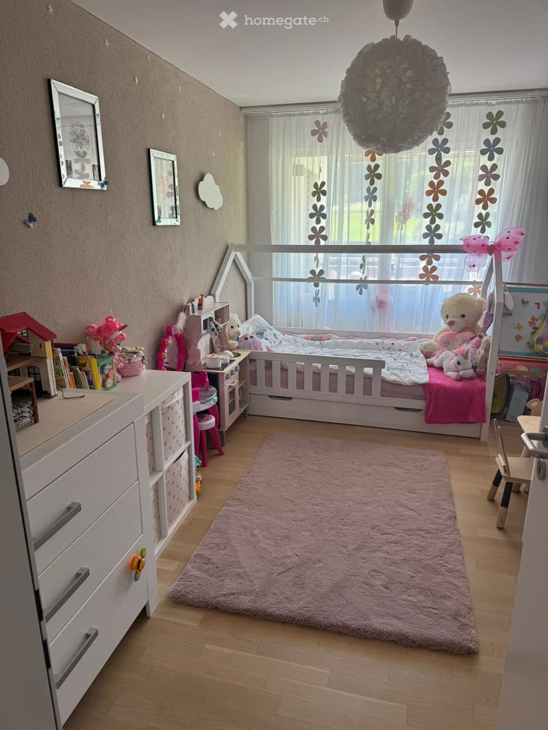 Cozy children's bedroom with a white wooden bed, pink bedding, a large pink rug, a nightstand, a small table, and various toys and decorations.