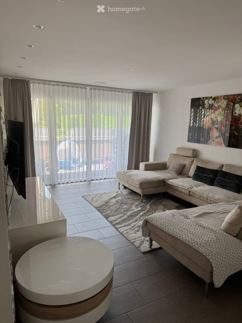 Modern living room with a large sectional sofa, coffee table, television, and artwork on the wall. The space features large windows with sheer curtains and a view of a balcony.