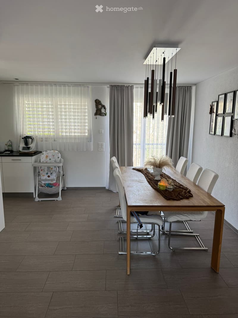 Dining room with a long wooden dining table, six white chairs, a baby chair, a kitchen appliance on a counter, and a chandelier hanging from the ceiling.