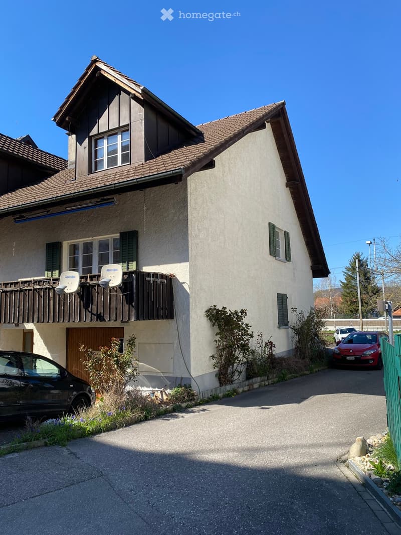 Two-story house with beige walls, tiled roof, and multiple windows. Features a balcony with satellite dishes, a garage, and a driveway with parked cars.