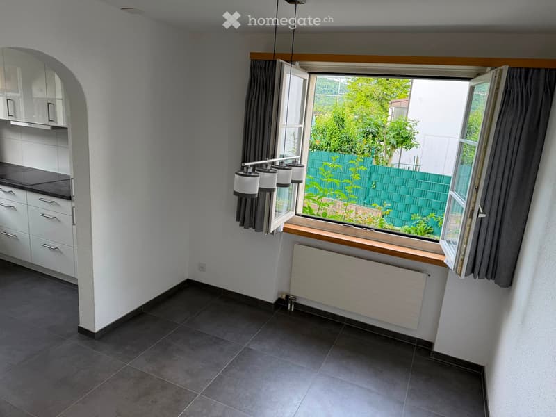 Modern kitchen with white cabinets, dark countertops, and a large window overlooking a garden. Features include a radiator under the window and a hanging light fixture.