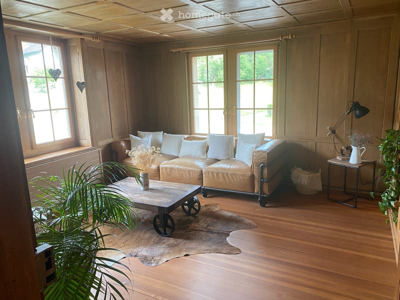 Cozy living room with wooden floors, large windows, a leather sofa, coffee table, side table, and plants.