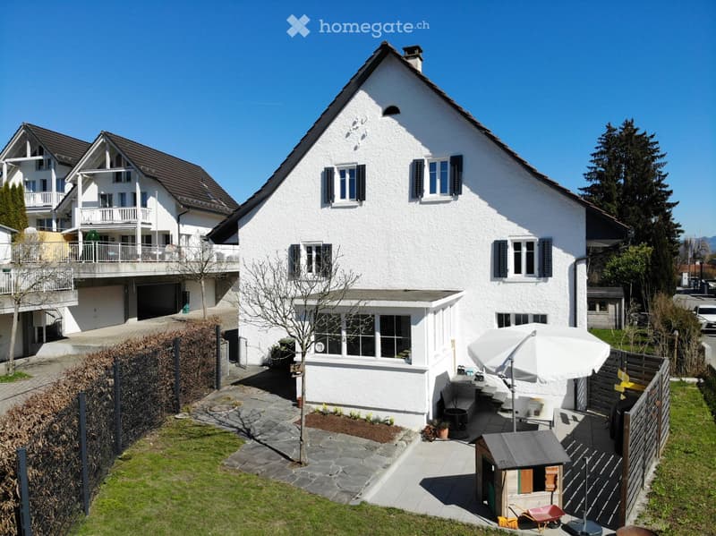 Two story house, white walls, brown roof, multiple windows, garden, patio, shed