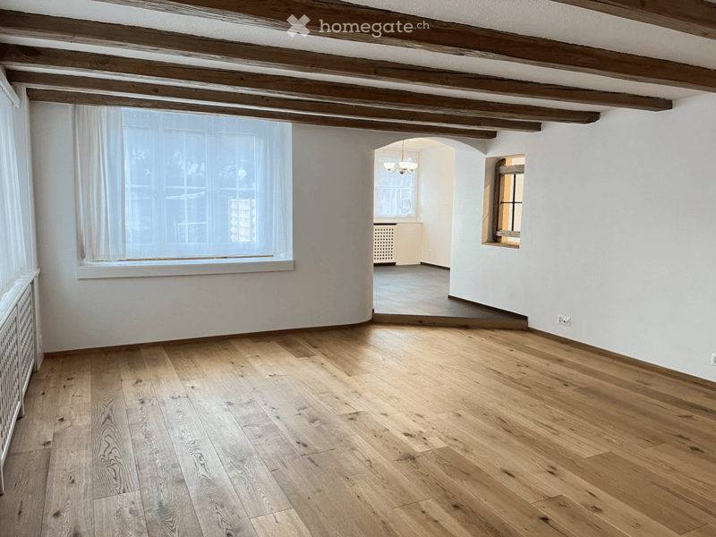 Hardwood floors, large window with white curtains, wooden ceiling beams, archway leading to another room, second small window, white walls, power outlets