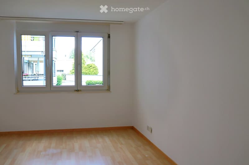 Empty room with light wooden flooring and white walls, featuring large windows with a view of an outdoor area.