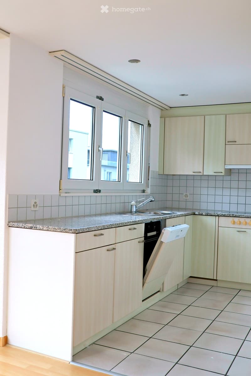 Modern kitchen with light wooden cabinets, granite countertop, sink, faucet, oven, and dishwasher. The space features tiled walls and a large window with blinds.
