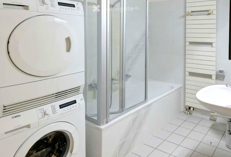 Modern bathroom with white tiled walls and floor, featuring a washing machine, a glass-enclosed shower, and a sink.