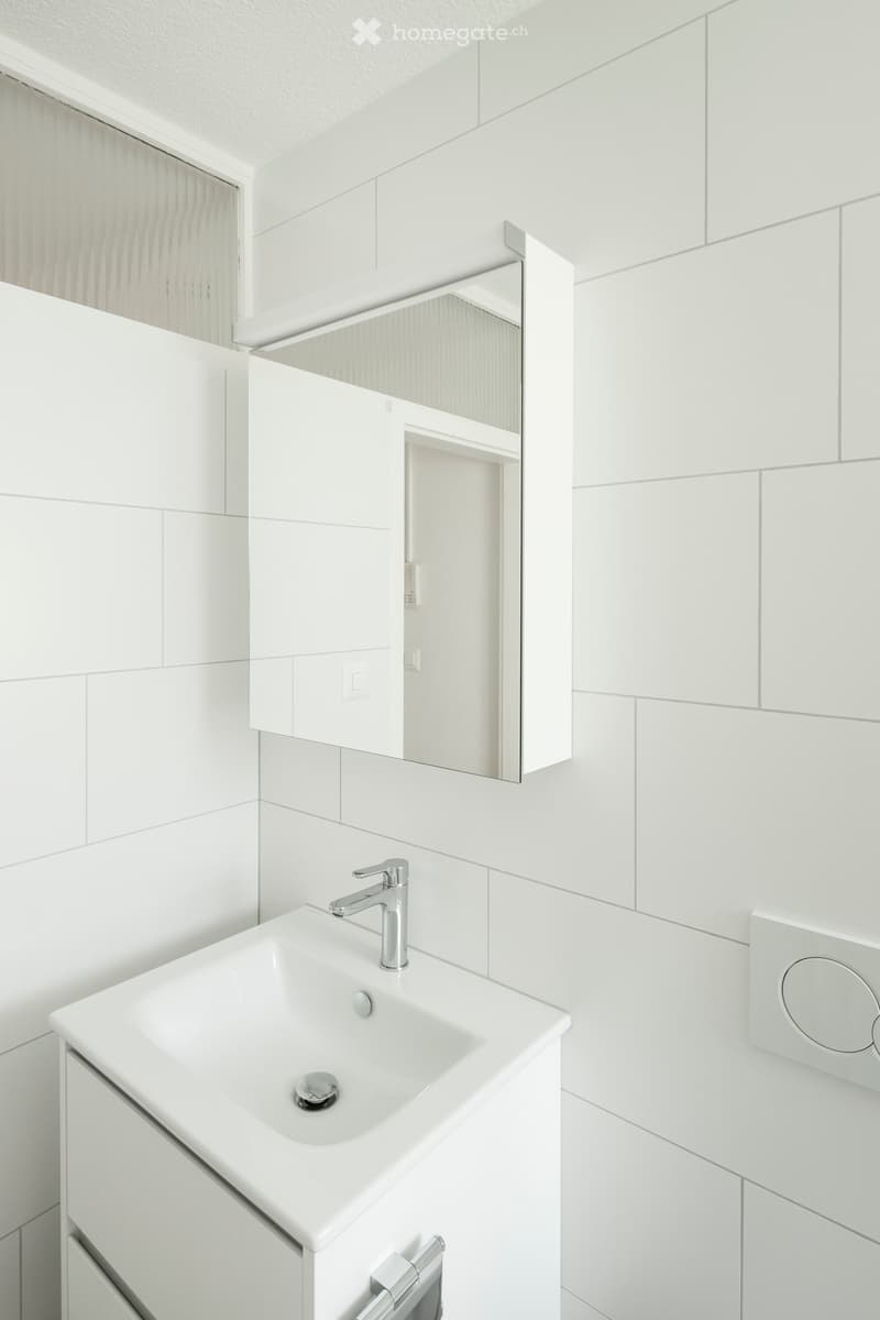 Modern bathroom with white tiles, a white sink, a chrome faucet, a mirrored cabinet, and a built-in storage unit.