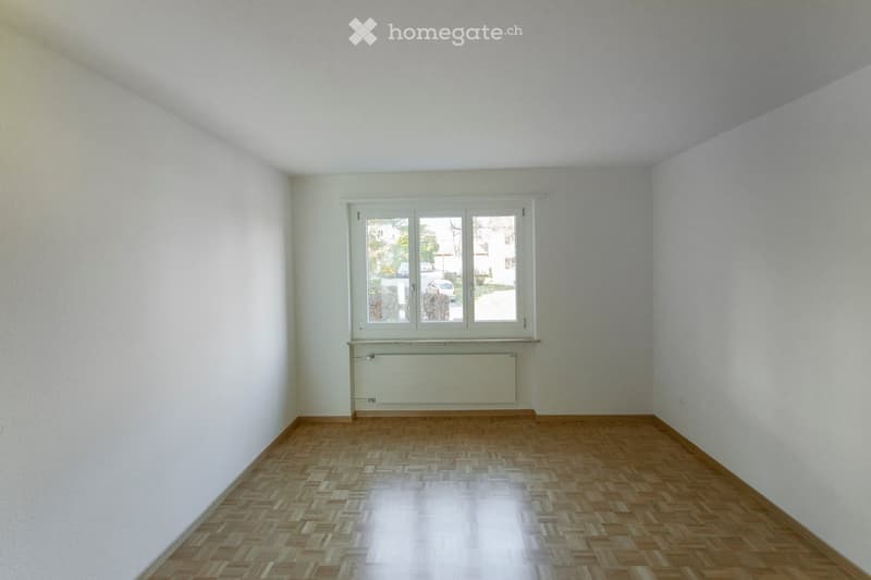 Empty room with light-colored walls, wooden parquet flooring, and a single window with white frame and radiator below it.