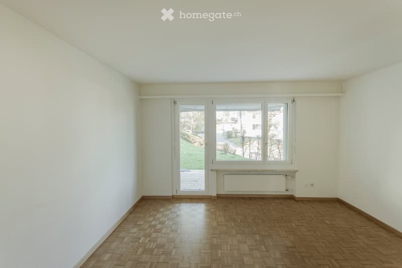 Empty room with light-colored walls, wooden floor, large windows, and a sliding glass door leading to an outdoor area.
