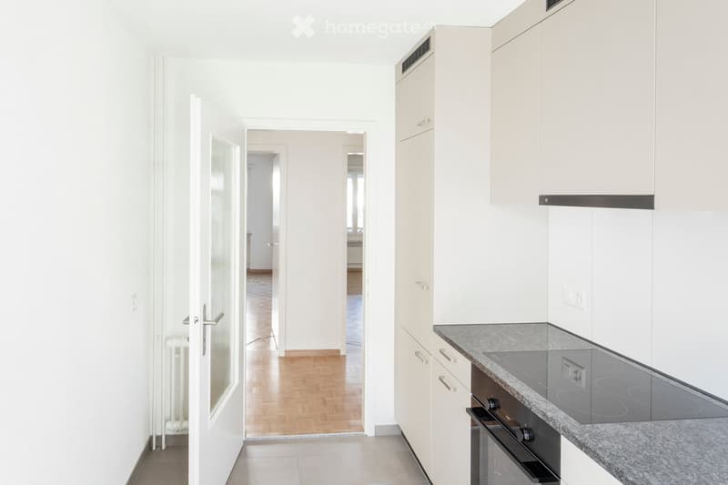 Modern kitchen with white cabinets, granite countertop, induction cooktop, and built-in oven. The hallway leads to another room with wooden flooring.