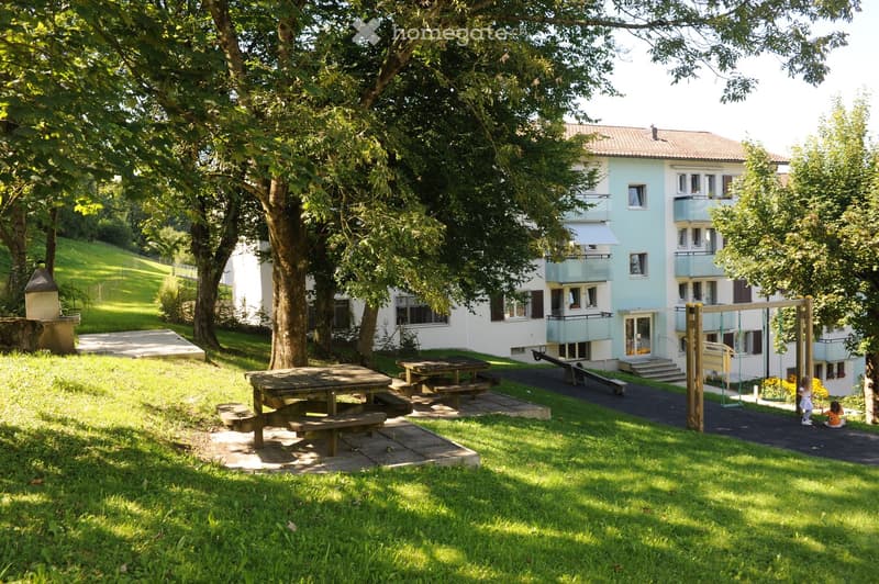 A well-maintained garden area with a light blue building in the background, featuring picnic tables, a swing set, and a grassy lawn.