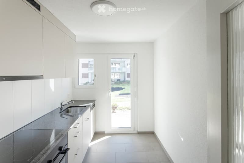 Modern, minimalist kitchen with white cabinets, stainless steel sink, and a large window overlooking a garden area.