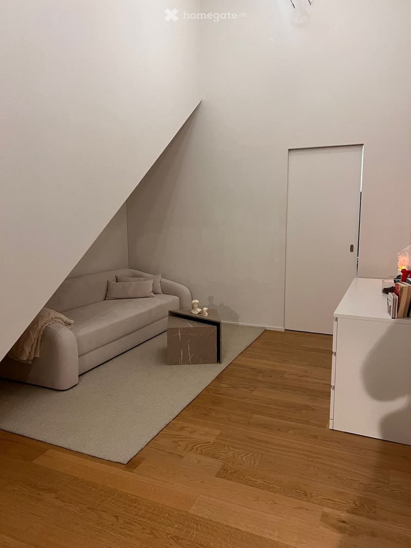 Modern living room with a light grey sofa, coffee table, side table, books, and a sculpture. The room features wooden flooring and a neutral color palette.