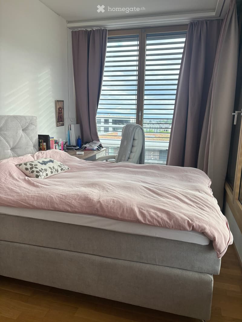 A modern bedroom with a large bed covered in pink bedding, a grey upholstered chair, a desk with books and a laptop, and a large window with blinds and curtains offering a view of the city.