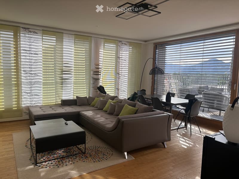 Modern living room with large windows offering a scenic mountain view, featuring a grey sectional sofa, coffee table, dining area with chairs, and a floor lamp.