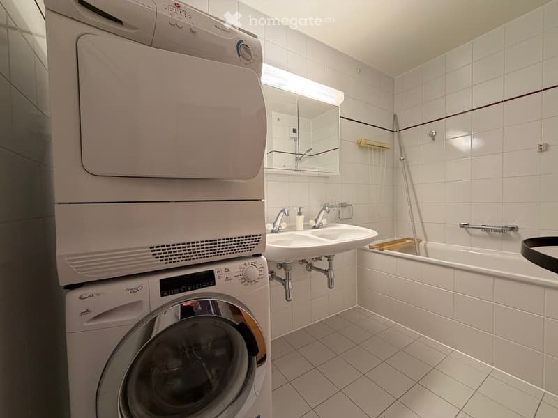 Modern bathroom with white tiled walls and floor, featuring a washing machine and dryer unit, a double sink, a bathtub, and a mirror above the sink.