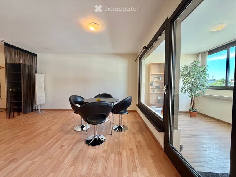 Modern dining area with wooden flooring, black chairs, round table, large windows offering a view, and a small shelf with a lamp.