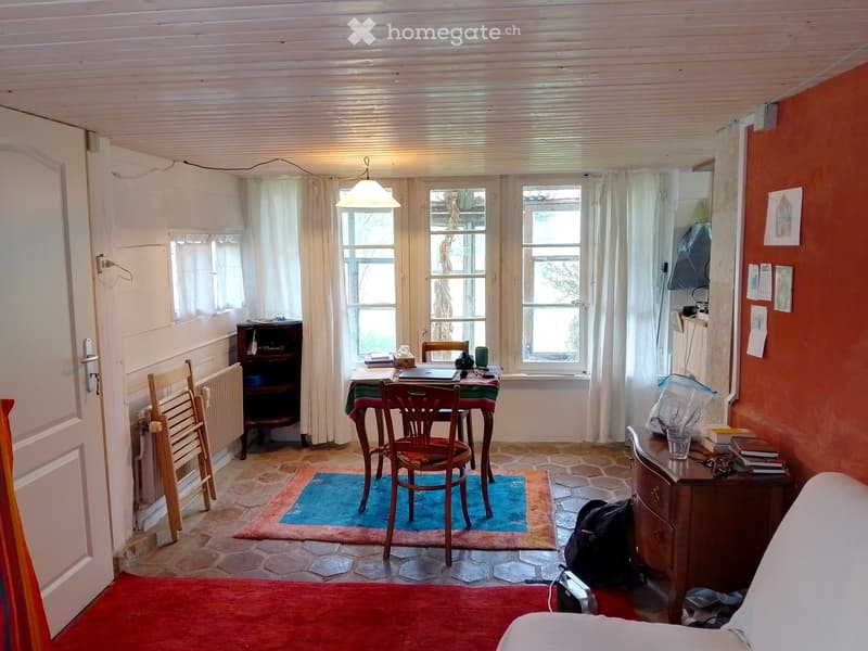 Cozy room with a bed, a small table with chairs, a laptop, a backpack, and a radiator. The room features a mix of red and blue rugs, a wooden cabinet, and a window with white curtains.