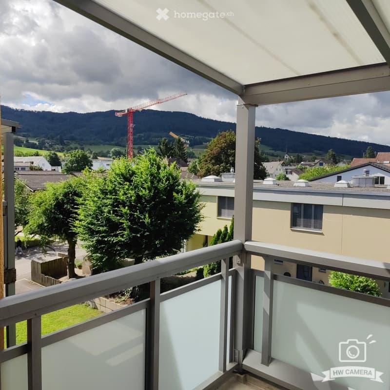Balcony with a view of a green landscape, trees, and distant mountains. The balcony has a modern metal railing and a covered section.