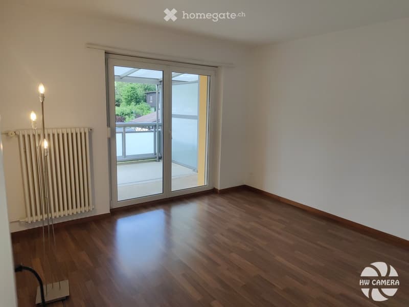 Empty room with wooden flooring, white walls, large sliding glass doors leading to a balcony, and a radiator on one wall.