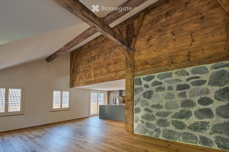 Open-concept space with wooden beams, exposed wooden ceiling, stone wall, and hardwood flooring. The area includes large windows and a view of a modern kitchen in the background.