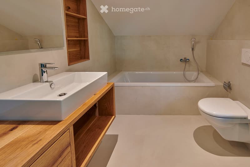 Modern bathroom with a white sink, wooden cabinet, bathtub, toilet, and a showerhead. The walls are light-colored, and there is a wooden shelf unit next to the sink.