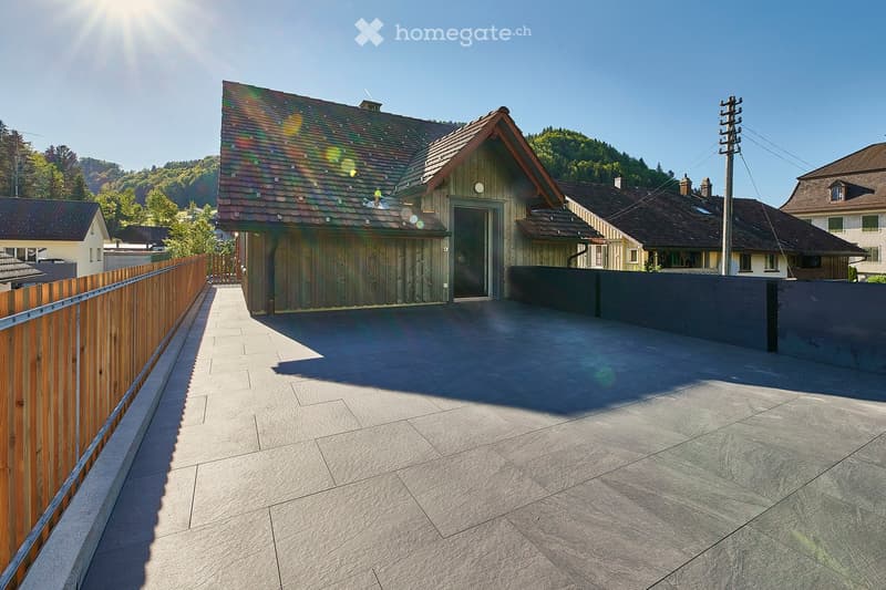 A spacious outdoor patio with a smooth concrete floor, bordered by a wooden fence on one side and a dark gray wall on the other. The area leads to a wooden house with a colorful roof and a small entrance door.