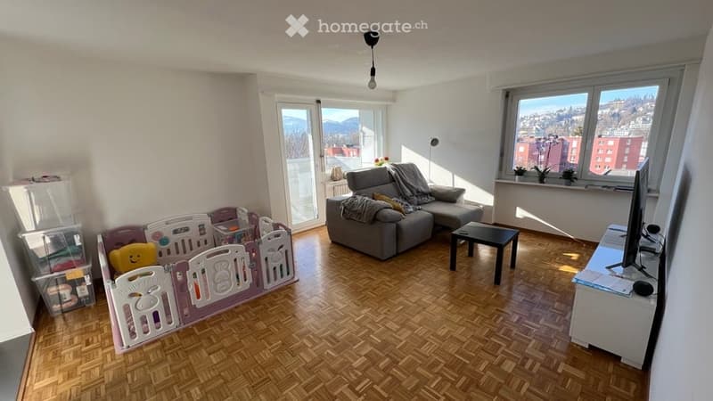 Bright living room with wooden flooring, large windows, and a view of the city and mountains. Features a grey sectional sofa, a small black coffee table, a TV stand, and a colorful playpen with a teddy bear.