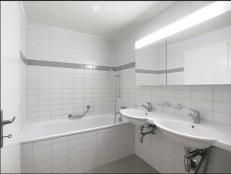 Modern bathroom with white tiled walls and floor, featuring a bathtub, two sinks with chrome faucets, a large mirror with built-in lighting, and a white cabinet.