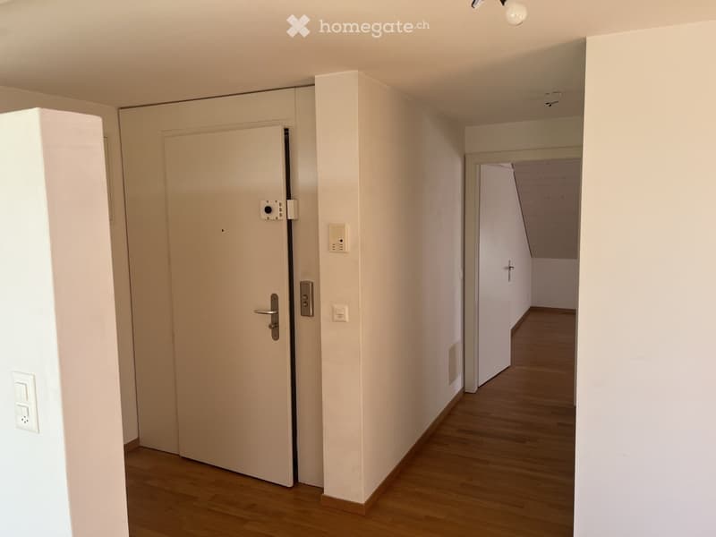 Empty hallway with wooden flooring, white walls, and a closed door on the left. A small section of another room is visible at the end of the hallway.